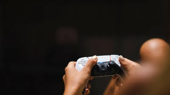 Player holding a white PS5 controller with both thumbs near the analog sticks in front of a TV