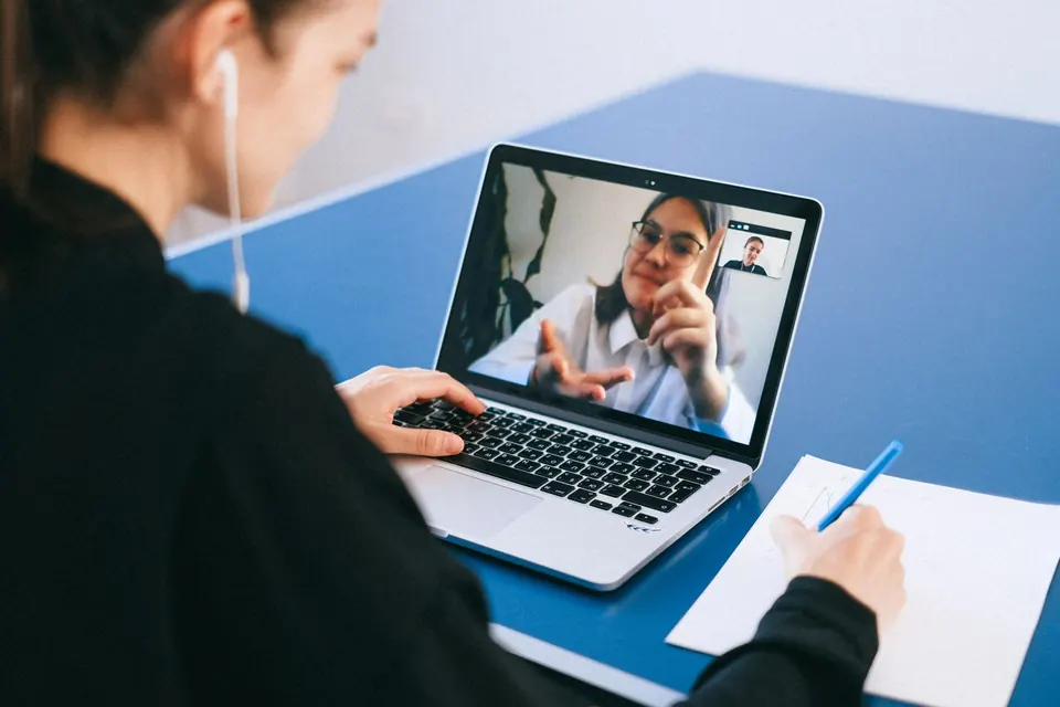 Person taking notes at a desk during a video call with a colleague on a laptop screen