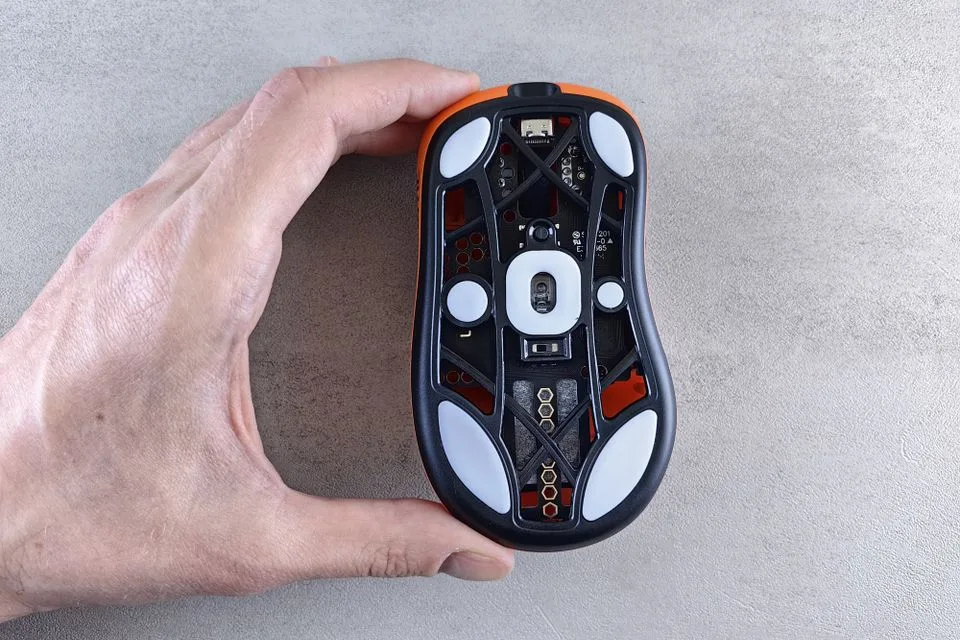 Hand holding a gaming mouse upside down showing the sensor opening and PTFE feet on its underside