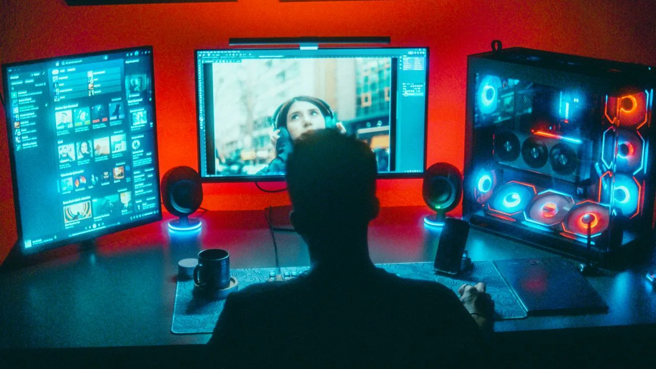 A gamer at a dark desk in front of a bright RGB monitor setup, the static-HUD, high-brightness usage pattern that most raises OLED burn-in risk