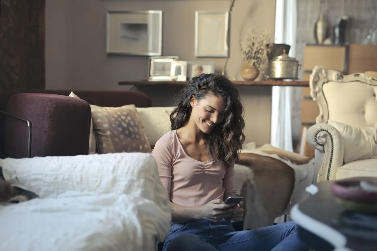 A person sitting in a bright living room holding a phone, the everyday setting where people ask how loud their room is and reach for an online decibel meter