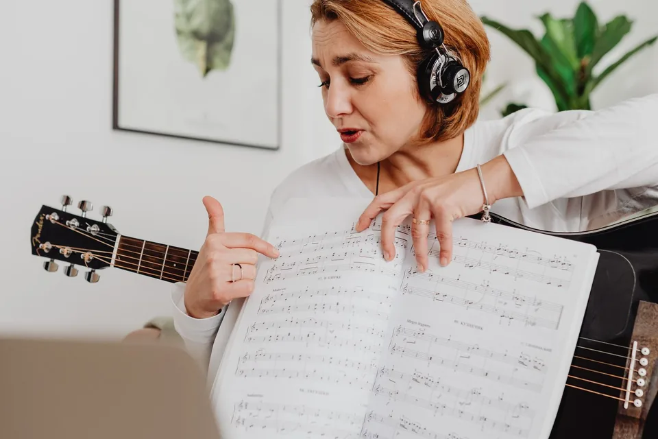 Singer with headphones practicing notes from sheet music at home with a guitar and a laptop nearby