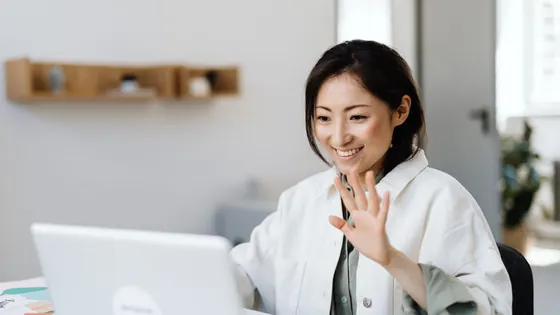 Smiling woman raising her hand in a wave at her laptop during a video call in a bright office