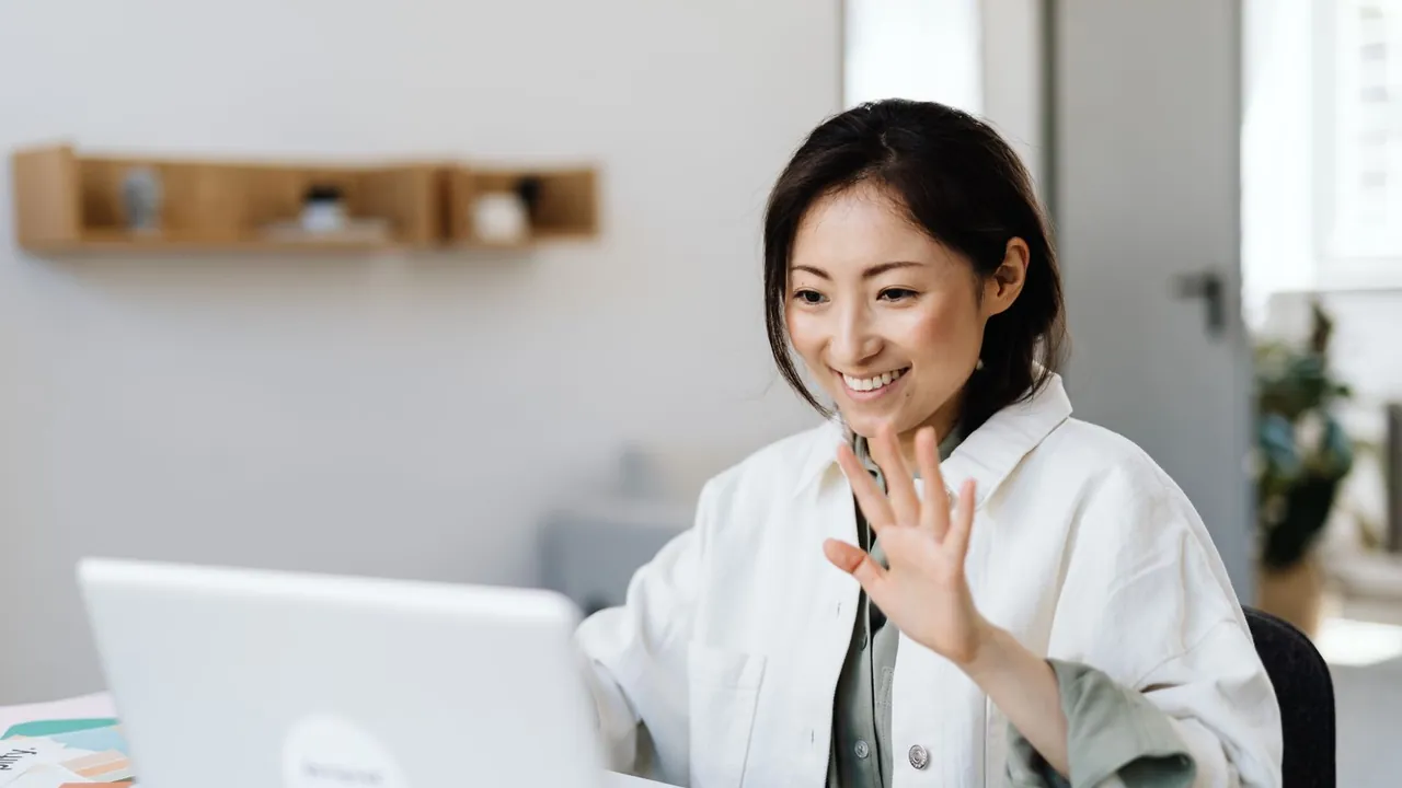 Smiling woman raising her hand in a wave at her laptop during a video call in a bright office