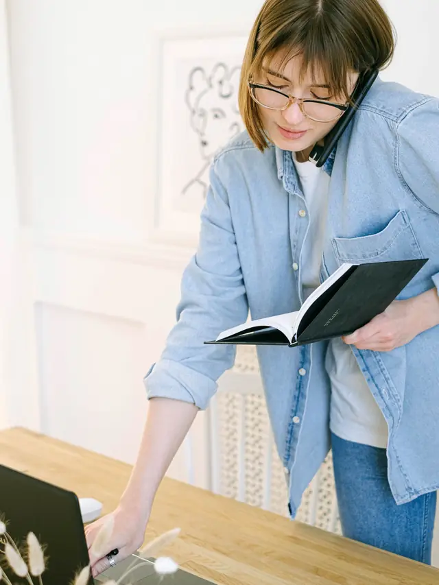 Woman on a phone call while checking notes beside a laptop
