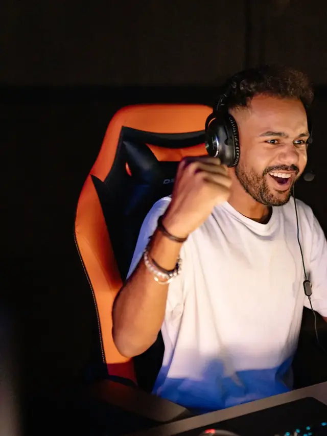 Excited esports player in a white T-shirt and headset clenching his fist at a monitor win in a dark gaming arena